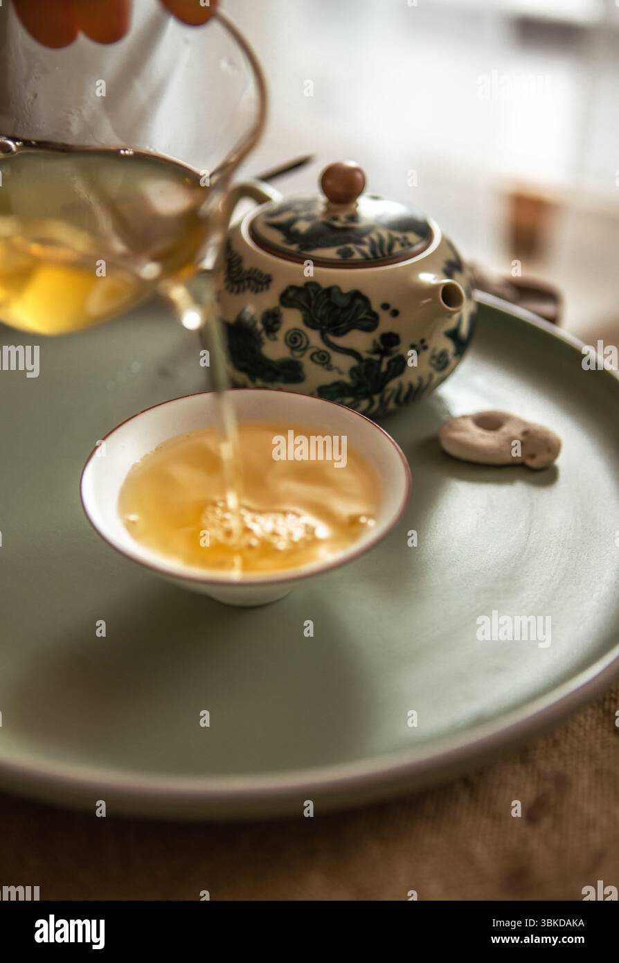 Hände, die Tee aus klarem Glaskrug in Teetassen gießen, schaffen ein ruhiges Teerritual, während sanftes Licht und hölzerner Teeteller an beruhigendes, achtsames Chinesisch erinnern Stockfoto