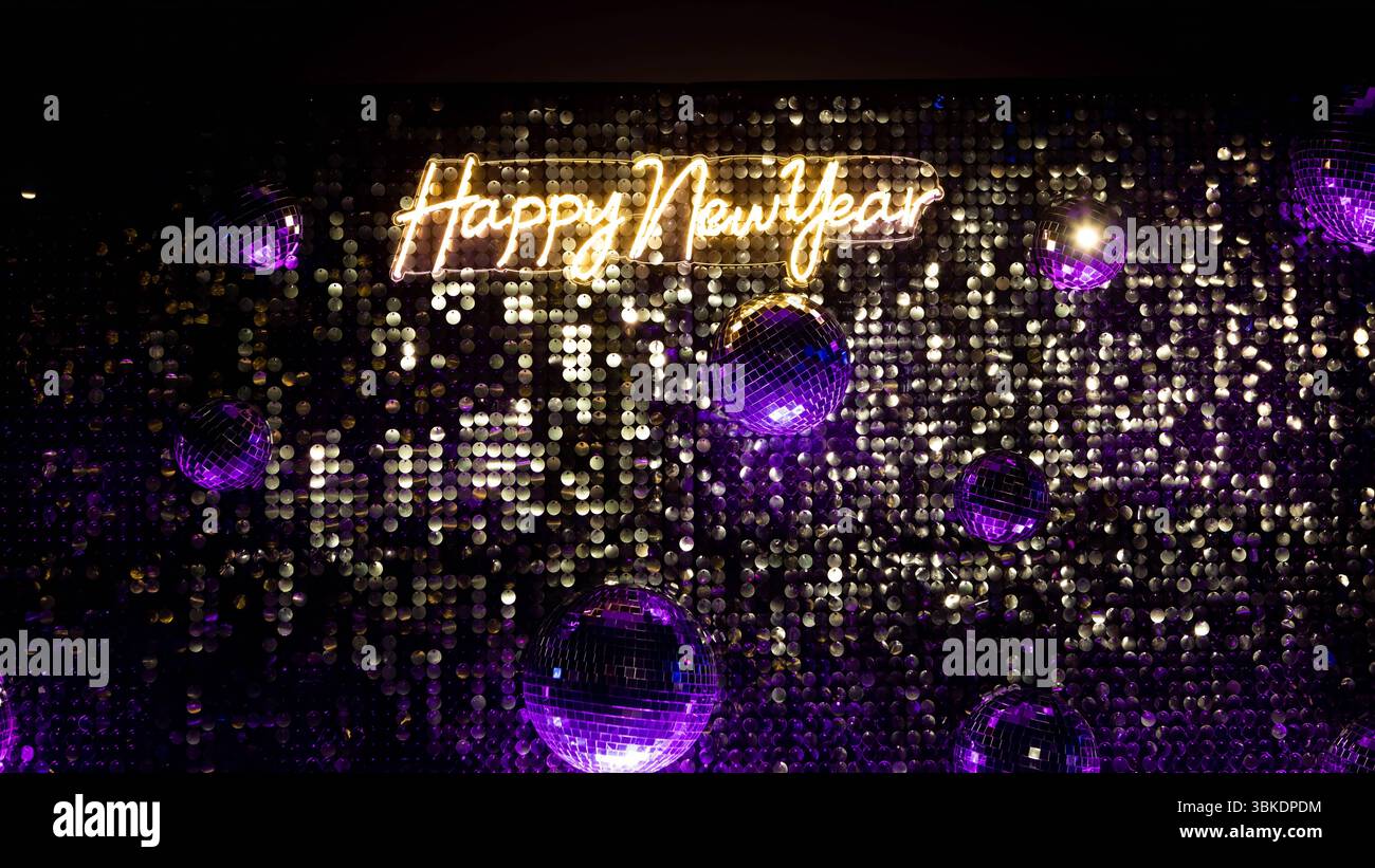 Eine Nahaufnahme einer glitzernden Pailletten-Kulisse mit Discokugeln, die ein leuchtendes Neonschild „Happy New Year“ hervorhebt. Stockfoto