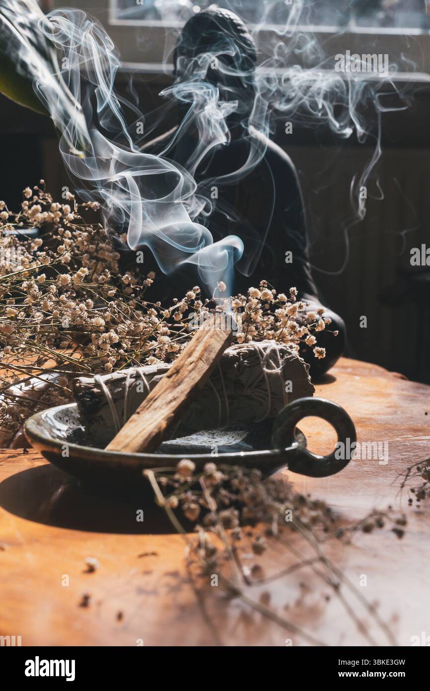 Brennender Salbei und Palo Santo kleben mit sich kräuselndem Rauch, umgeben von getrockneten Blumen auf Holztisch Stockfoto