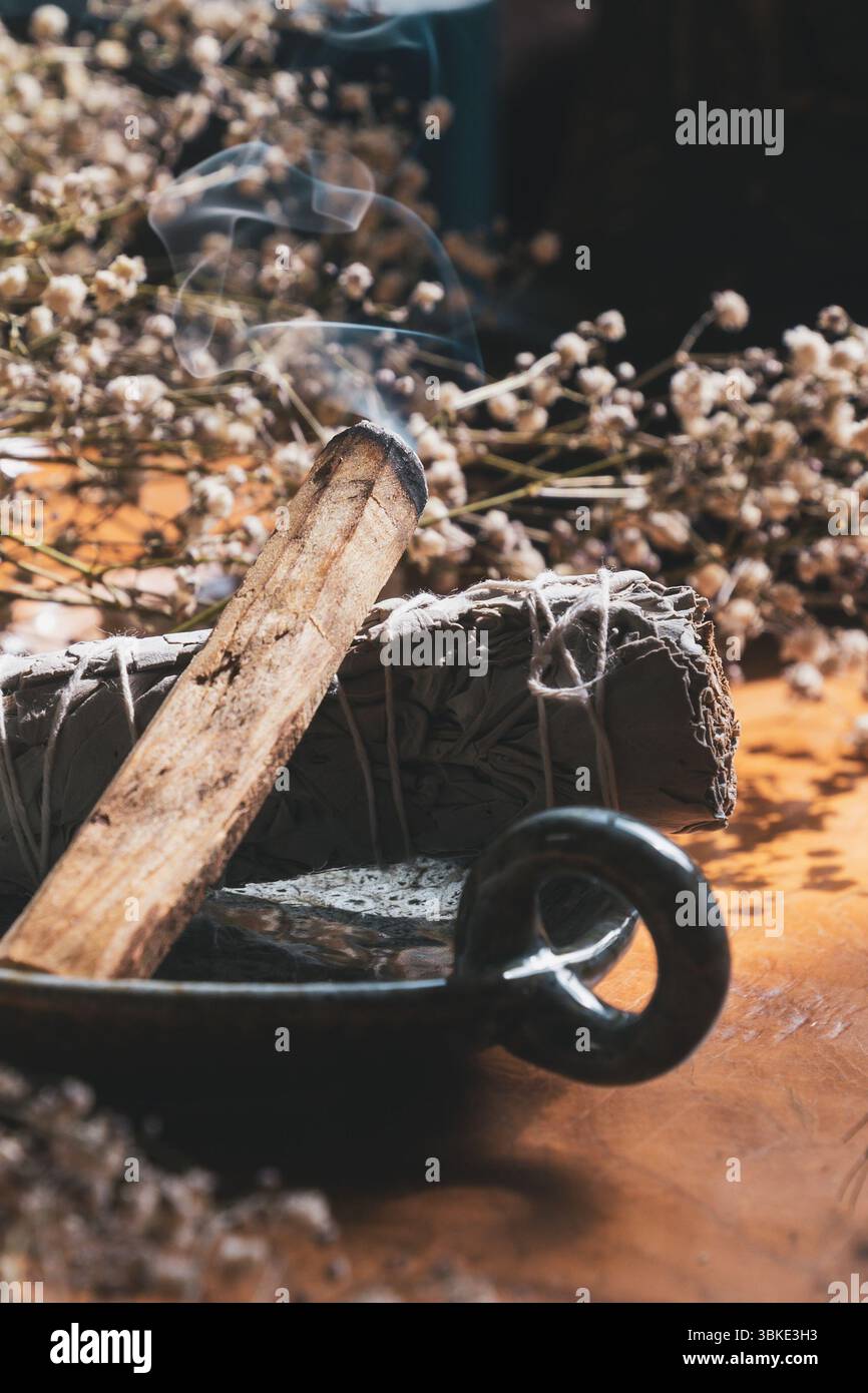 Nahaufnahme von brennendem Palo Santo und Salbeischmierstab mit sichtbarem Rauch, umgeben von getrockneten Blumen Stockfoto