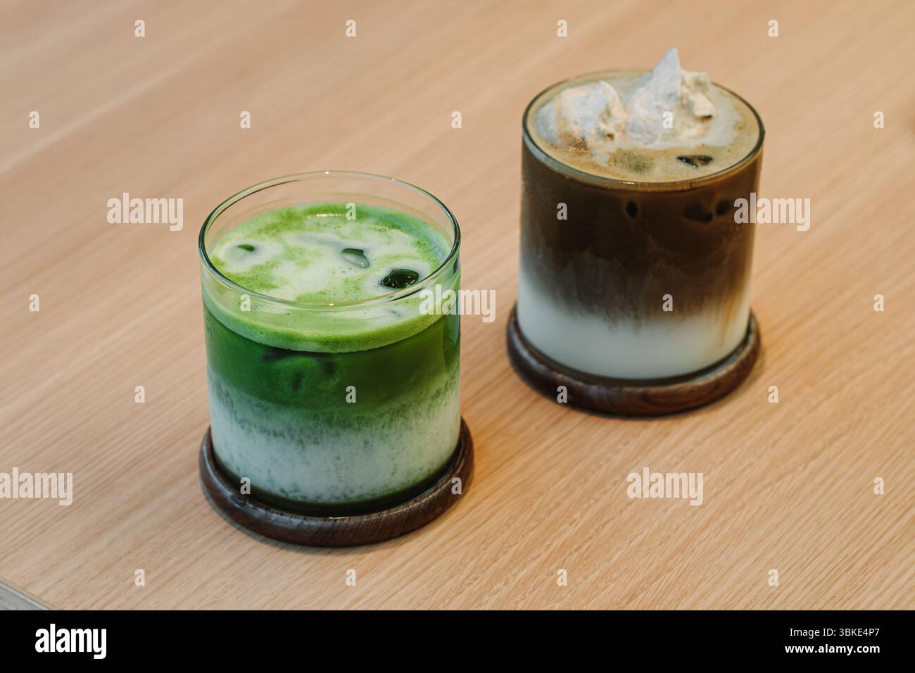 Zwei Eisgetränke, Matcha Latte und Hojicha Latte, serviert in klaren Glasbechern auf hölzernen Untersetzern auf einem hellen Holztisch Stockfoto
