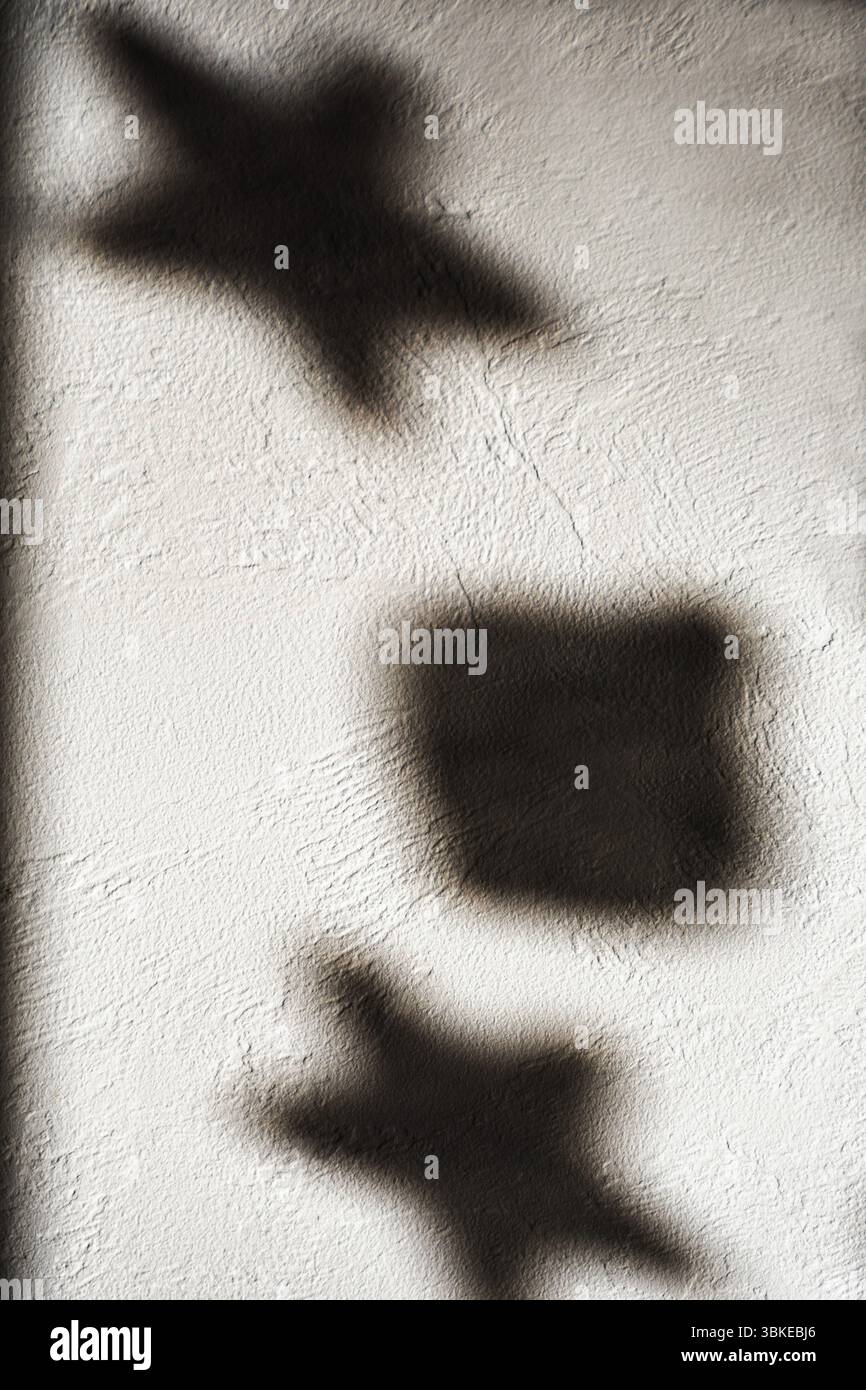 Die Formen von Sternen und ein Quadrat werfen Schatten auf eine strukturierte weiße Wand, die von sanftem Licht beleuchtet wird. Stockfoto