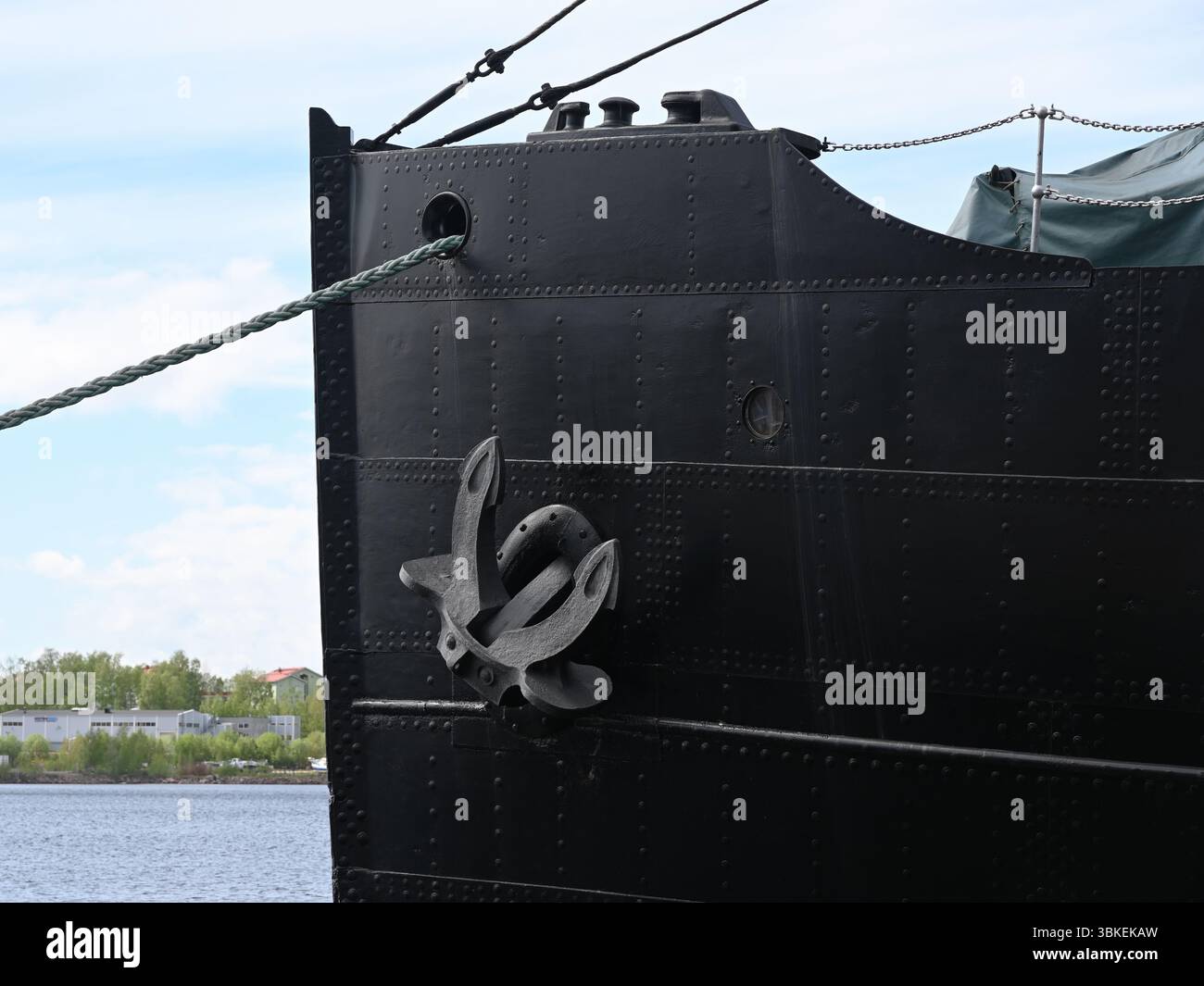 Der imposante Bug eines großen schwarzen Schiffes, der an einem Hafen ankert, mit einem markanten Stahlanker, der an seinem genieteten Metallrumpf befestigt ist. Das industrielle Design Stockfoto