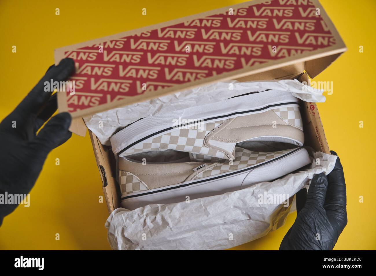 Mit Handschuhen öffnen Sie einen Schuhkarton für Vans, der neue karierte Slip-on-Schuhe vor einem leuchtend gelben Hintergrund enthüllt Stockfoto