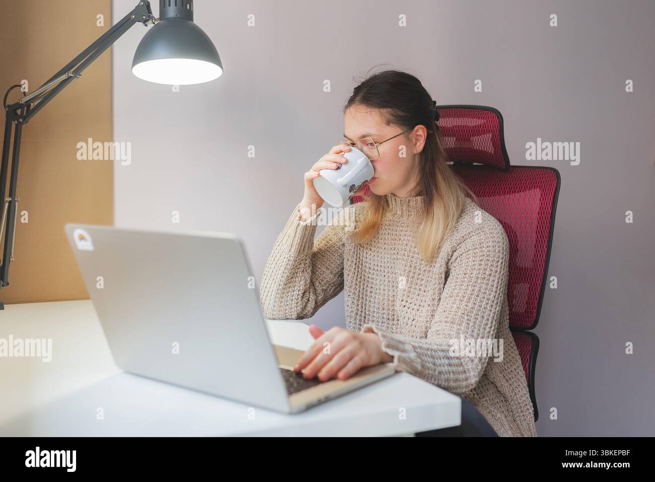 Frau trinkt aus einem Becher, während sie von zu Hause aus auf einem Laptop arbeitet. Roter Mesh-Stuhl, beigefarbener Strickpullover und konzentrierter Blick auf den Bildschirm, während sie aus weißem M schlürft Stockfoto