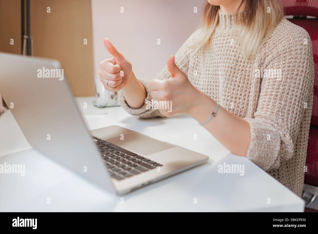 Frau gibt während eines positiven Online-Meetings am Schreibtisch zwei Daumen hoch. Lächelnde Frau zeigt zwei Daumen nach oben vor dem Laptop, legere und warme Kleidung, Stockfoto