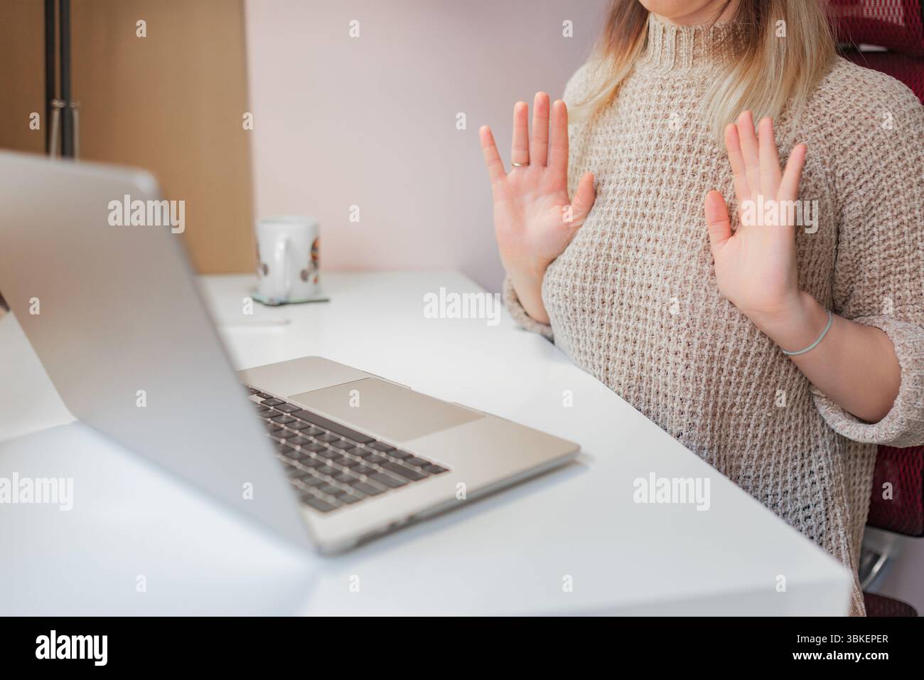 Frau hebt während des Online-Meetings in neutraler Geste beide Hände leicht. Die Hände sind entspannt hoch, die Frau sitzt während der virtuellen Sitzung am Schreibtisch, leicht Stockfoto