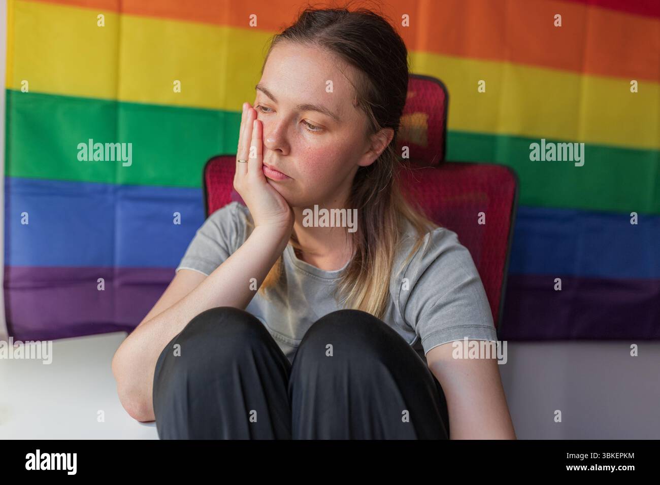 Eine nachdenkliche Frau, die Kopf auf der Hand liegt, vor dem Hintergrund des Regenbogenstolzes sitzt. Die Frau sitzt mit gezogenen Knien, das Gesicht auf der Hand liegend, nachdenkliche Expensionen Stockfoto