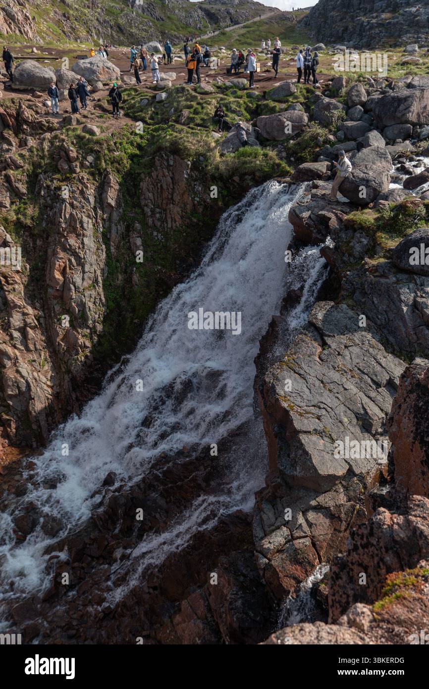 Teriberka, Russland - 13. Juni 2025: Touristen versammeln sich an einem sonnigen Tag um den Batarayskiy-Wasserfall in Teriberka, umgeben von schroffen arktischen Felsen Stockfoto