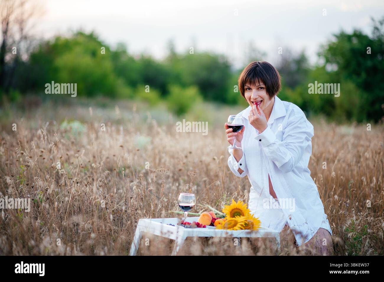 Brünette vor dem Hintergrund eines trockenen Feldes. Lächelndes Mädchen mit einem Glas Wein im Urlaub. Obst, Blumen und Wein, um Ihre Stimmung zu heben. Stockfoto