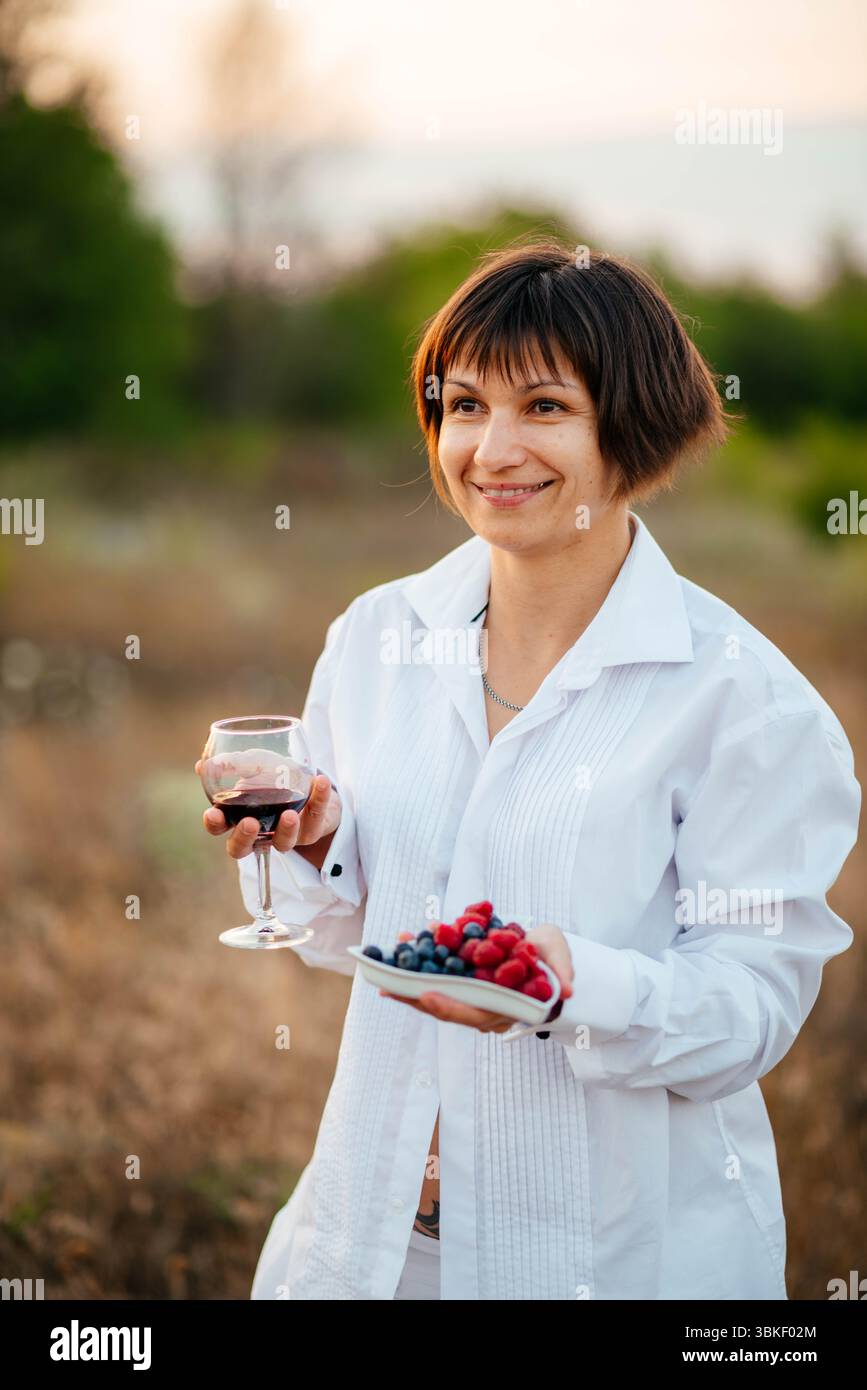 Brünette vor dem Hintergrund eines trockenen Feldes. Lächelndes Mädchen mit einem Glas Wein im Urlaub. Obst, Blumen und Wein, um Ihre Stimmung zu heben. Stockfoto