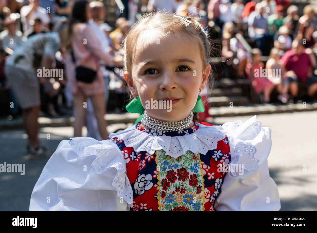 Porträt eines Mädchens in traditioneller Sliac Volkstracht mit Blick auf die Kamera, mit Leuten auf einer kleinen Tribüne im Hintergrund beim Juniales Folk Festival Stockfoto