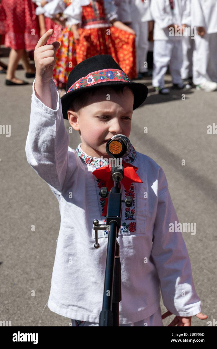 Selbstbewusster Junge in traditioneller Sliac Volkstracht, der beim Juniales Folk Festival mit erhobener Hand ins Mikrofon spricht Stockfoto