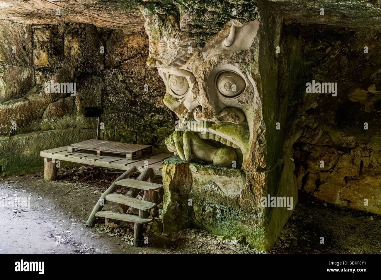 Die Lapidiales-Skulpturen in Port-d'Envaux, Nouvelle-Aquitaine, Frankreich, umfassen eine mythische Kreatur mit großen spiralförmigen Augen unter einem Felsvorsprung. Eine menschliche Figur steckt in ihrem Maul. Moos bedeckt Teile des Steins. Links führen Holzstufen zu einer Plattform aus Brettern hinauf. Verschiedene Künstler haben die Lapidiales in den Fels eines ehemaligen Steinbruchs gemeißelt. Die Werke stellen verschiedene Figuren und Szenen dar. Die Kunstwerke sind Teil der Landschaft und fügen sich harmonisch in die natürliche Umgebung ein. Sie sind ein Beispiel für die Kunst und Geschichte der Region. Der Bildhauer Alain Tenenbaum gründete th Stockfoto