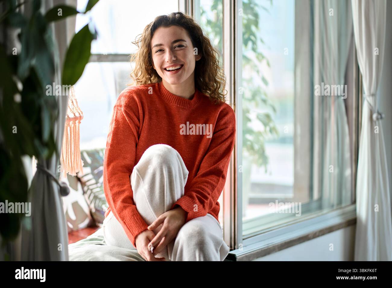 Glückliche, hübsche junge Frau, die zu Hause auf dem Fensterbrett sitzt und lacht. Stockfoto