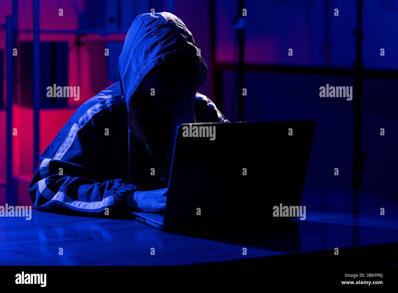 Ein Mann in einem schwarzen Kapuzenpullover, der in einem neonbeleuchteten Raum einen Laptop benutzt und sein Gesicht im Schatten versteckt hat. Er erinnert an Themen wie Cybersicherheit, Hacking, Codin Stockfoto