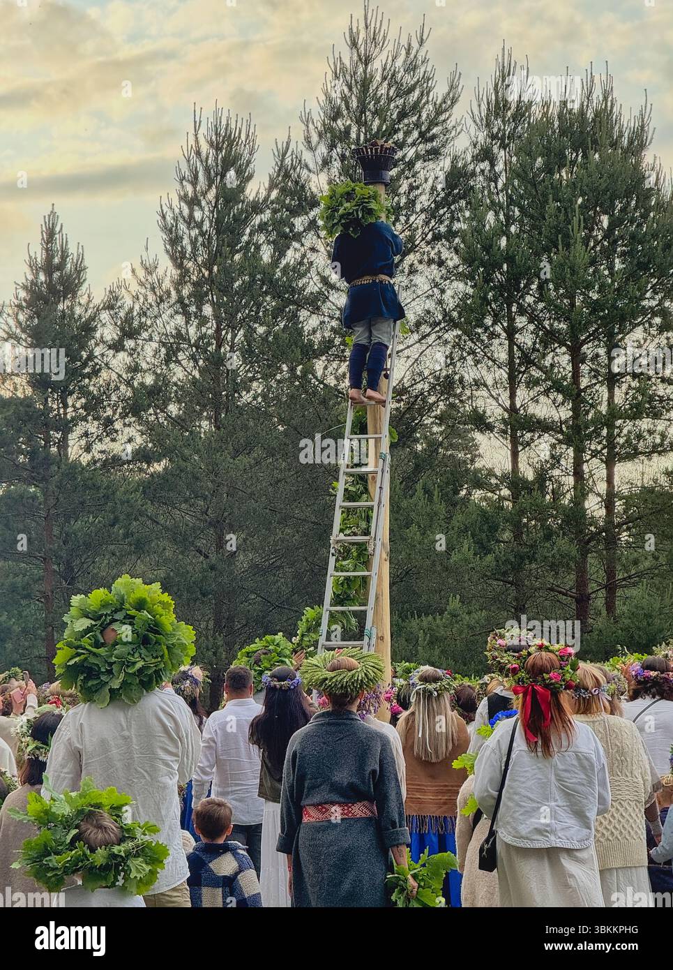 Menschen in traditionellen lettischen Trachten und Kränzen beobachten, wie ältere Menschen im Hochsommer lange Kränze an der Stange befestigen Stockfoto