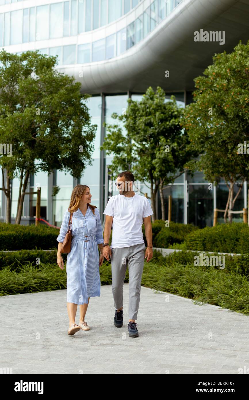Ein Mann und eine Frau gehen Hand in Hand durch einen üppigen Stadtpark, umgeben von Grün und modernen Gebäuden, und genießen ihre gemeinsame Zeit. Stockfoto