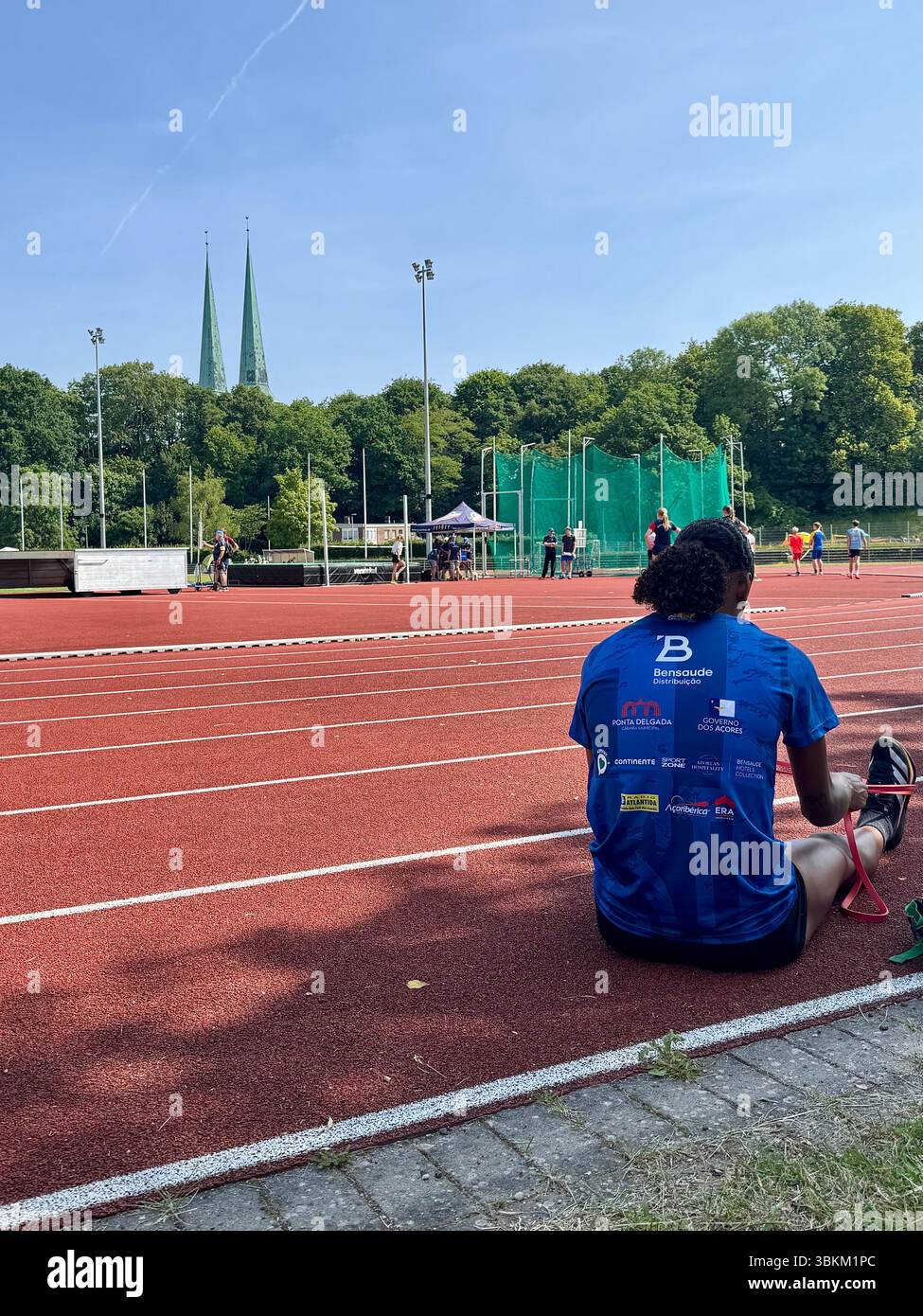 Der Athlet sitzt an einem sonnigen Tag in Lübeck am Rande einer Leichtathletikstrecke, mit Kirchtürmen und Bäumen, die sich auf den Wettkampf vorbereiten - Smartphone-aufgenommenes Stockfoto
