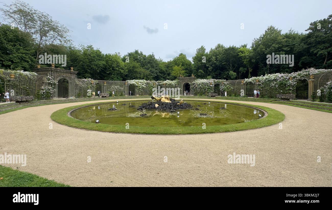 Oben ein Rundteich mit einer Goldstatue in der Mitte, umgeben von Spalierbögen, die mit weißen Blumen und grünen Bäumen bedeckt sind. Stockfoto