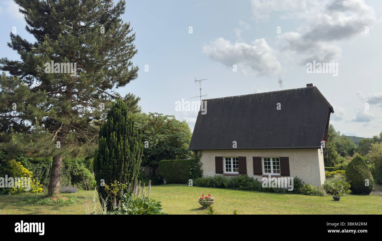 Blick von außen auf ein malerisches beigefarbenes Haus mit dunklem Dach und gepflegtem Rasen, umgeben von üppigem Grün und einer großen Kiefer. Stockfoto