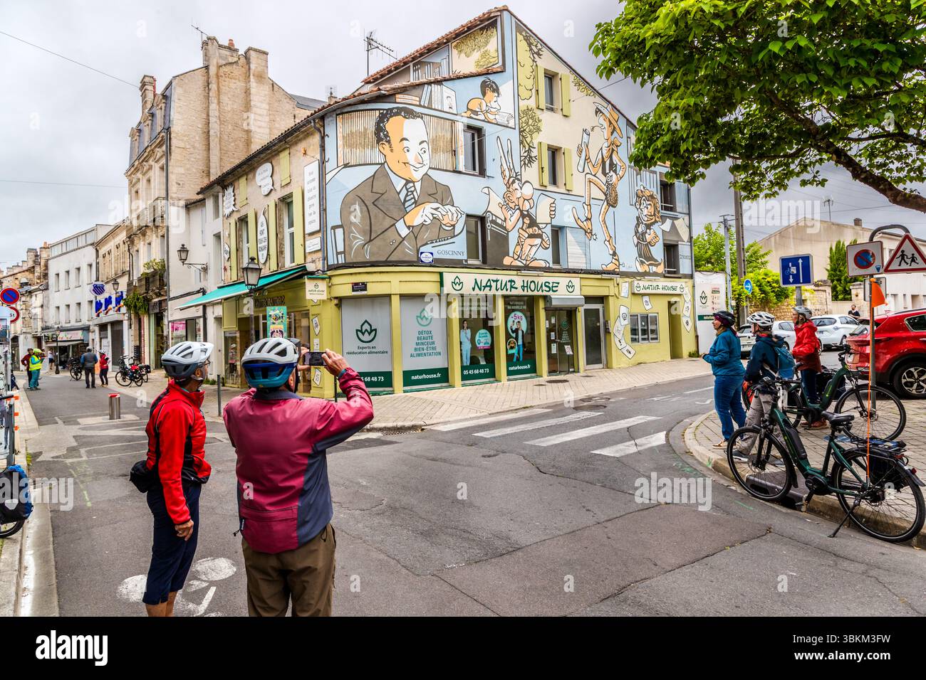 Radtouristen in Angoulême vor dem Wandbild „die Welt von René Goscinny“, das den Karikaturisten mit seinen berühmtesten Figuren wie Asterix, Lucky Luke und Petit Nicolas zeigt. Eine Gruppe von Radfahrern hält an, um ein Wandgemälde anzuschauen und zu fotografieren, das die Fassade eines Gebäudes ziert. Das Wandbild zeigt Szenen aus den Comicbüchern von René Goscinny, einem berühmten französischen Comic-Autor, der in Angoulême geboren wurde. Die Szene fängt die lebhafte Atmosphäre der Stadt ein, die für ihre Comickunst berühmt ist. Angoulême ist seit 2019 als UNESCO-Kreativstadt in der Kategorie Literatur anerkannt. Rue Ren Stockfoto
