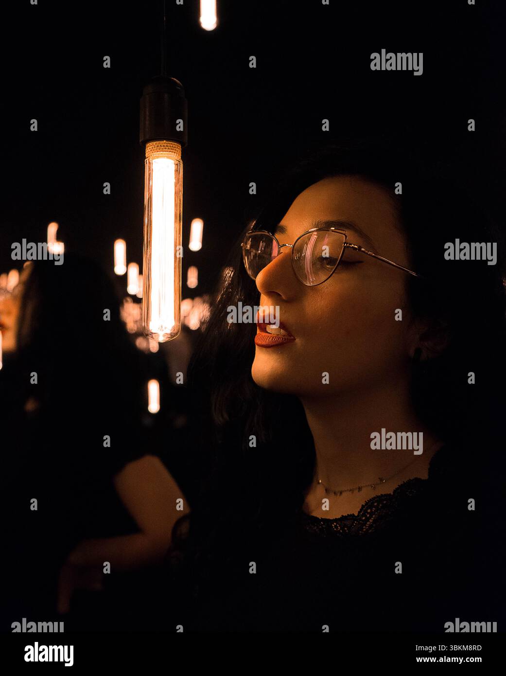 Eine Frau mit Brille schaut auf eine hängende Glühbirne in einem schwach beleuchteten Raum. Die Atmosphäre ist friedlich und ruhig. Stockfoto