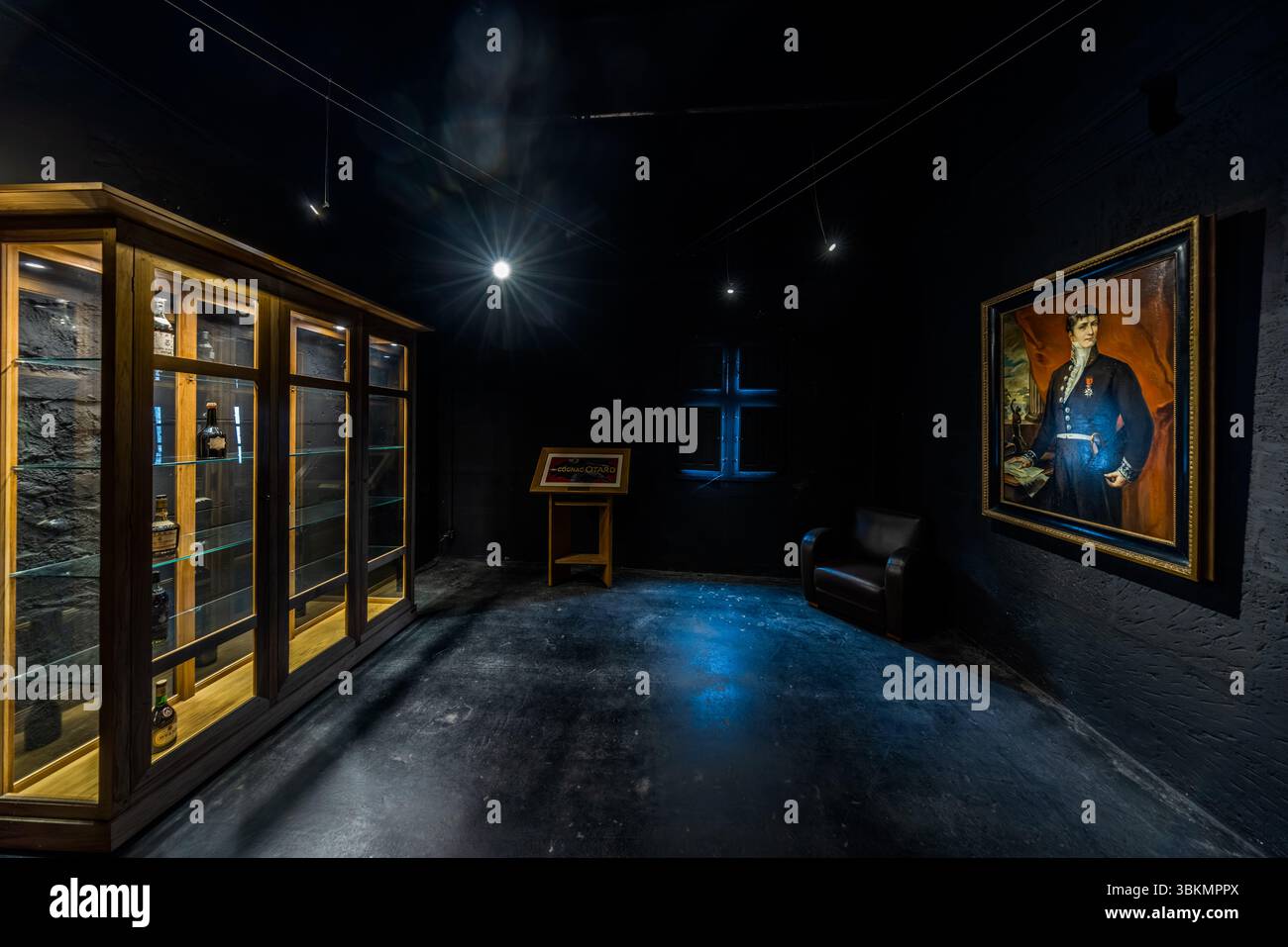 Ein abgedunkelter Ausstellungsraum des Hauses Baron Otard im Cognac-Schloss (Château de Cognac), Rue François 1er, Cognac, Nouvelle-Aquitaine, Frankreich, zeigt Cognac-Flaschen in einer beleuchteten Holzvitrine mit Glastüren. Rechts hält ein vergoldeter Rahmen das Porträt eines Mannes in einer dunklen bestickten Jacke mit einer Verzierung an einem roten Band (möglicherweise der Firmengründer Jean-Baptiste Antoine Otard). Er stützt eine Hand auf Papiere auf einem Tisch. In der Mitte stehen ein Lesepult mit einer Plakette und ein schwarzer Ledersessel. Ein Kreuzfenster leuchtet blau in der dunklen Wand. Jean-Baptiste Antoine Otard gründete t Stockfoto