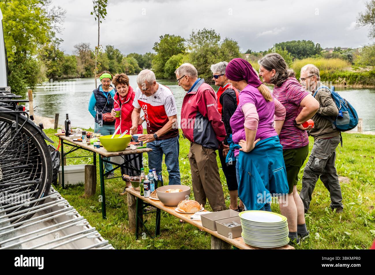 Teilnehmer an der geführten Radtour, organisiert von Die Landpartie, machen ein Picknick am Ufer der Charente in Le Ponton, Rouffiac, Nouvelle-Aquitaine, Frankreich. Auf halber Strecke der Etappe des Tages stärken sie sich mit regionalen Spezialitäten aus dem Departement Charente. Neun Personen stehen an zwei Tischen mit einer Salatschüssel, Brot, Getränken und einem Stapel Teller, wobei die Tourführerin Katell Nicoux mit einem grünen Kopfband ganz links steht. Das Unterstützungsfahrzeug mit einem Fahrradträger steht links geparkt. Die Gruppe fährt entlang der Charente auf der La Flow Vélo Fernradroute. Das Unterstützungsfahrzeug Stockfoto