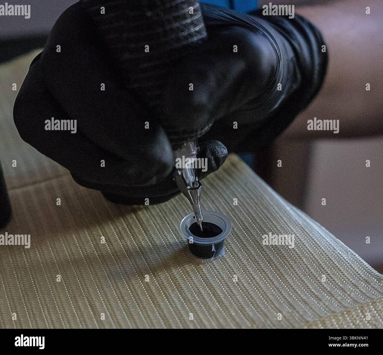 Eine Hand mit Handschuhen hält eine Tätowiermaschine mit einer Nadel und taucht sie in einen kleinen Behälter, der mit dunkler Tätowierfarbe gefüllt ist. Stockfoto