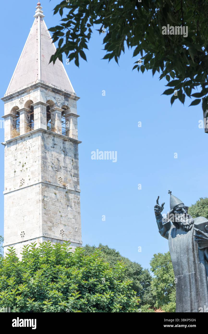 SPLIT, KROATIEN, 08.06.2018: Eine ruhige Szene mit einem historischen Glockenturm neben einer hohen Statue, umgeben von üppigem Grün, unter einem klaren blauen Himmel, Stockfoto