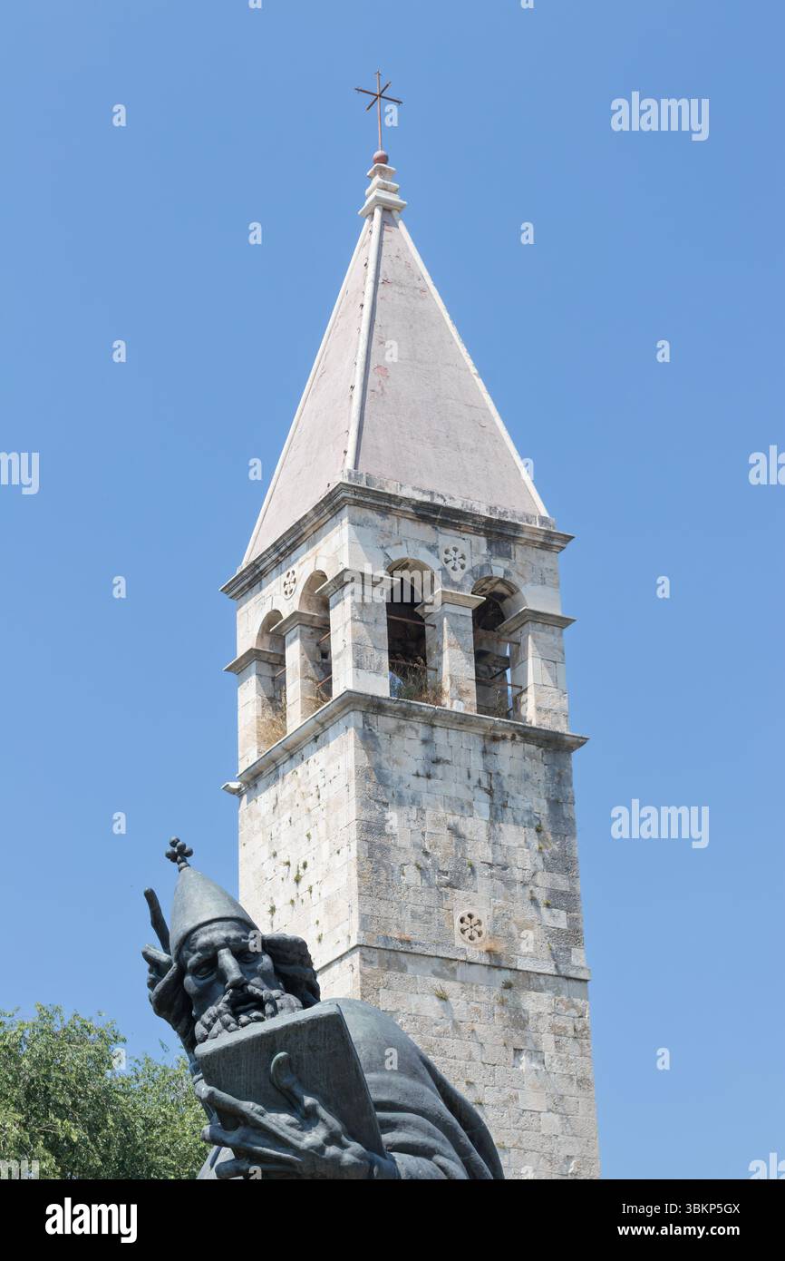 SPLIT, KROATIEN, 08.06.2018: Eine ruhige Szene mit einem historischen Glockenturm neben einer hohen Statue, umgeben von üppigem Grün, unter einem klaren blauen Himmel, Stockfoto