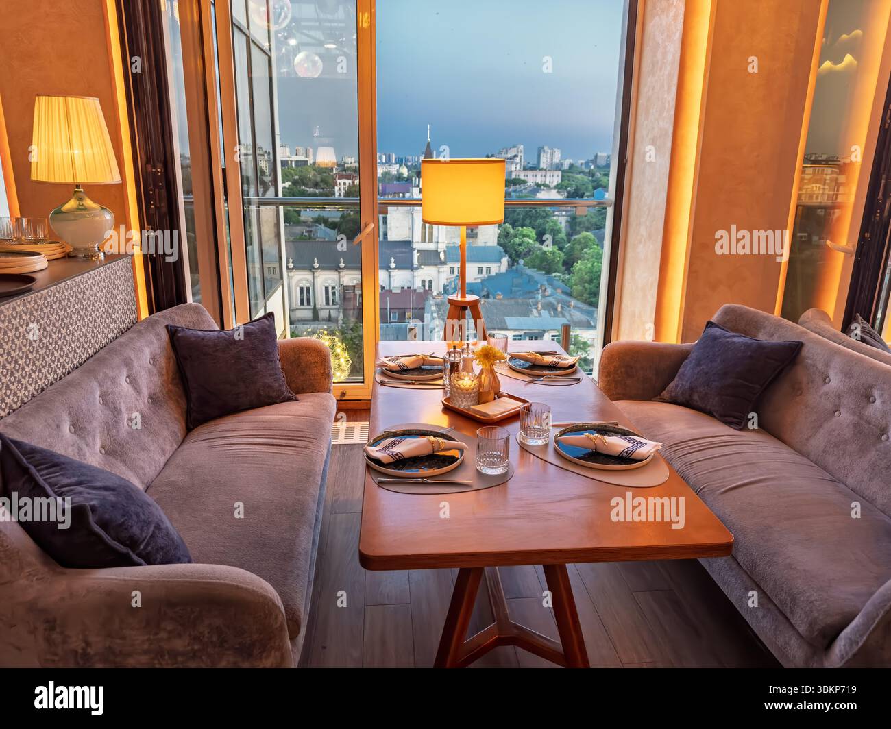 Wunderschön gestalteter Esstisch am Fenster in einem schicken Restaurant, mit warmem Umgebungslicht und den Lichtern der abendlichen Skyline im Stockfoto