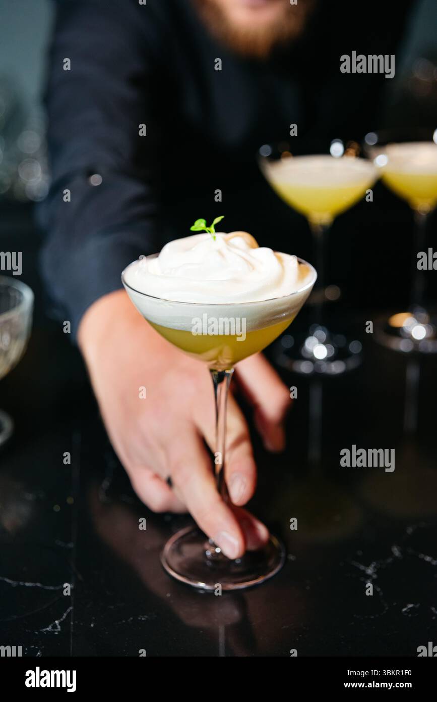 Nahaufnahme des Barkepers, der einen Zitruscocktail mit dickem Schaum und Kräutergarnitur in einem stilvollen Coupé-Glas präsentiert. Stockfoto