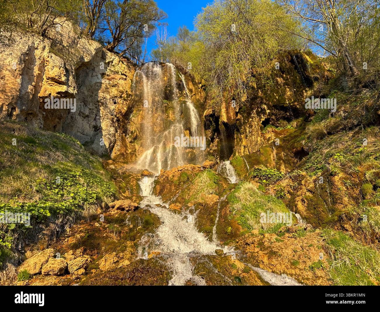 Wunderschöner Wasserfall, der eine felsige Klippe hinunterstürzt, umgeben von üppiger grüner Vegetation unter klarem blauem Himmel, gefangen in lebendigem Frühlingssonnenlicht. - Smartphone-aufgenommenes Stockfoto