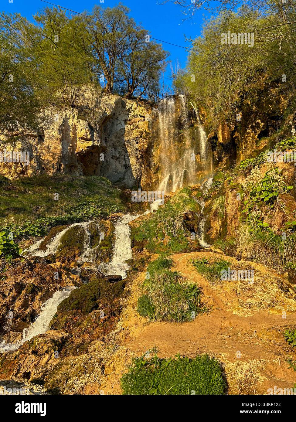 Ein malerischer Wasserfall, der über moosige Felsen stürzt, umgeben von üppigem Grün und Klippen unter einem klaren blauen Himmel, gefangen im warmen Sonnenlicht. - Smartphone-aufgenommenes Stockfoto