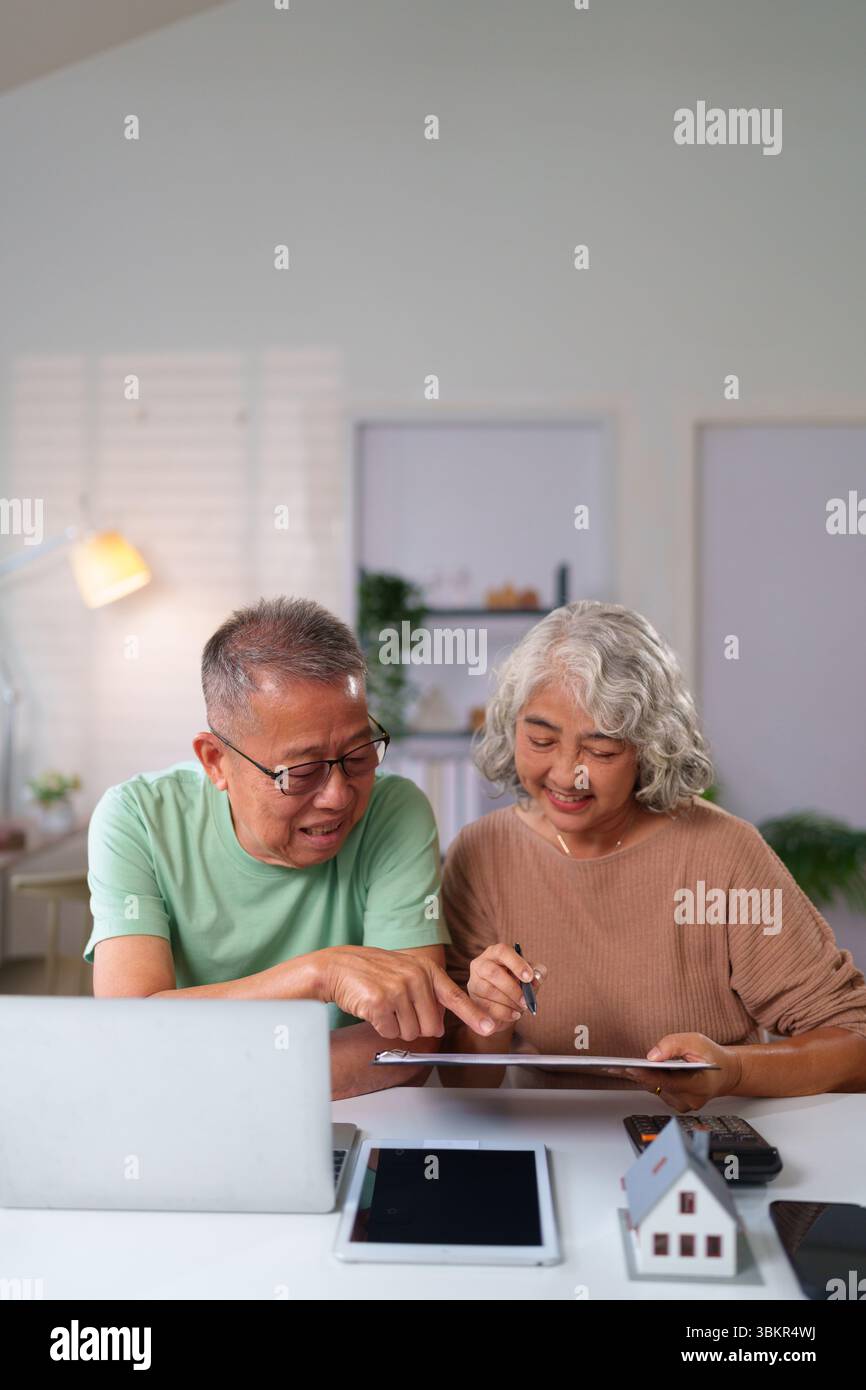 Ein älteres asiatisches Ehepaar prüft gerne die Finanzdokumente zu Hause mit einem Laptop, Tablet und Rechner, die für zukünftige Immobilieninvestitionen planen Stockfoto