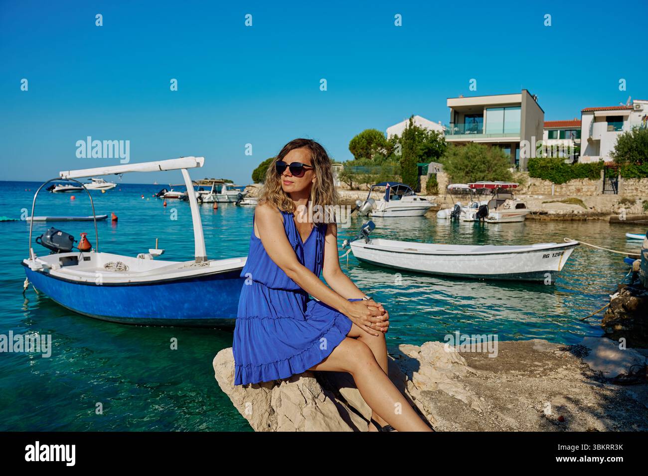 Frau in blauem Kleid sitzt auf Felsen in der Nähe des Meeres mit Booten auf türkisfarbenem Wasser. Sommerurlaub und Küstenleben Konzept Stockfoto