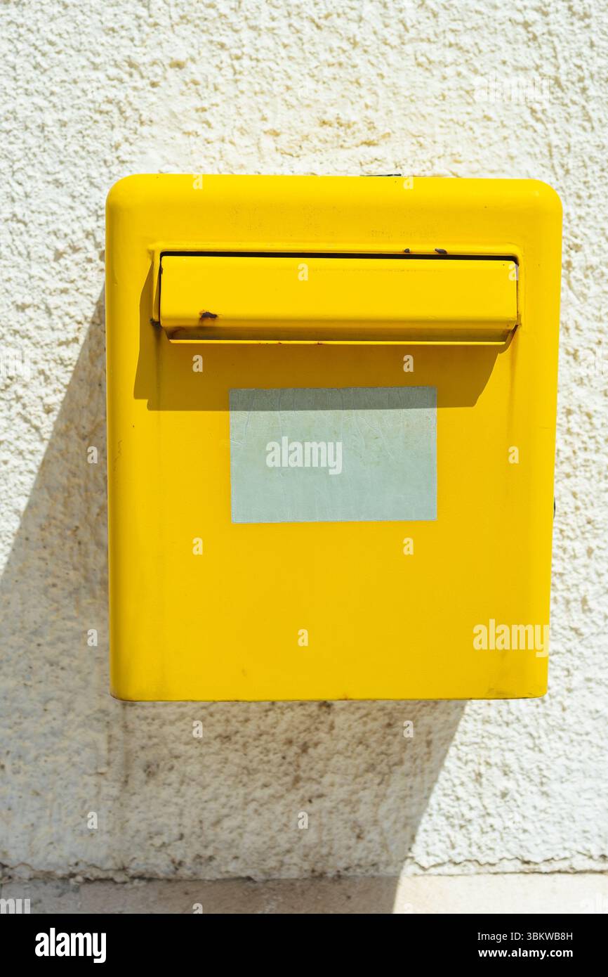Gelbe Mailbox an hellweißer Wand. Postdienst, Kommunikationssymbol und Straßendetails in sonniger Umgebung. Stockfoto