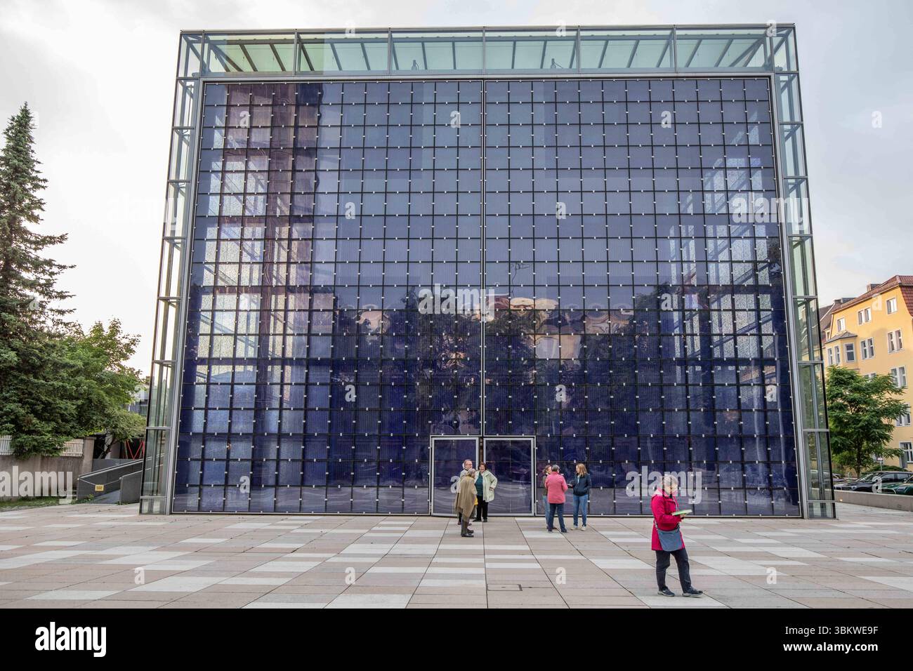 München, Deutschland 08. Juni 2025: Im Bild: Das Bild zeigt die markante Glasfassade der modernen Herz-Jesu-Kirche in München, Deutschland, die durch ihre innovative Architektur besticht. Die gesamte Front des Kirchengebäudes besteht aus einer großflächigen, in einzelne Quadrate unterteilten Blauglasfassade, die Bauwerk ein einzigartiges und transparentes Erscheinungsbild verleiht. Die Glaspaneele reflektieren die Umgebung und schaffen interessante Spiegelungen von Bäumen und dem Himmel. Im unteren Bereich sind die Eingangstüren erkennbar, vor denen sich eine weitläufige, höllisch gepflasterte Stockfoto