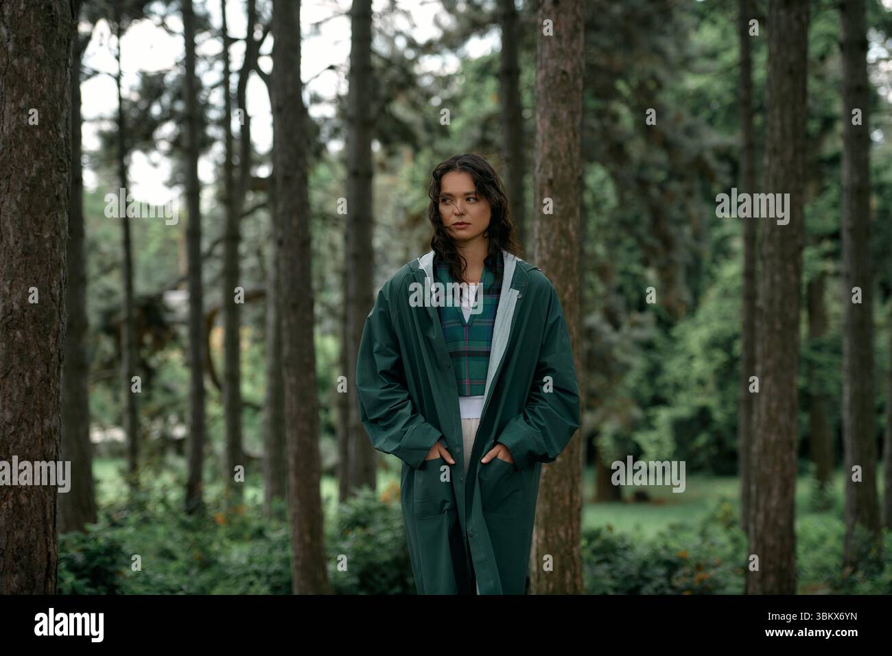 Eine junge Frau mit lockigen Haaren trägt einen trendigen Regenmantel, während sie im Sommer üppiges Grün entdeckt. Stockfoto