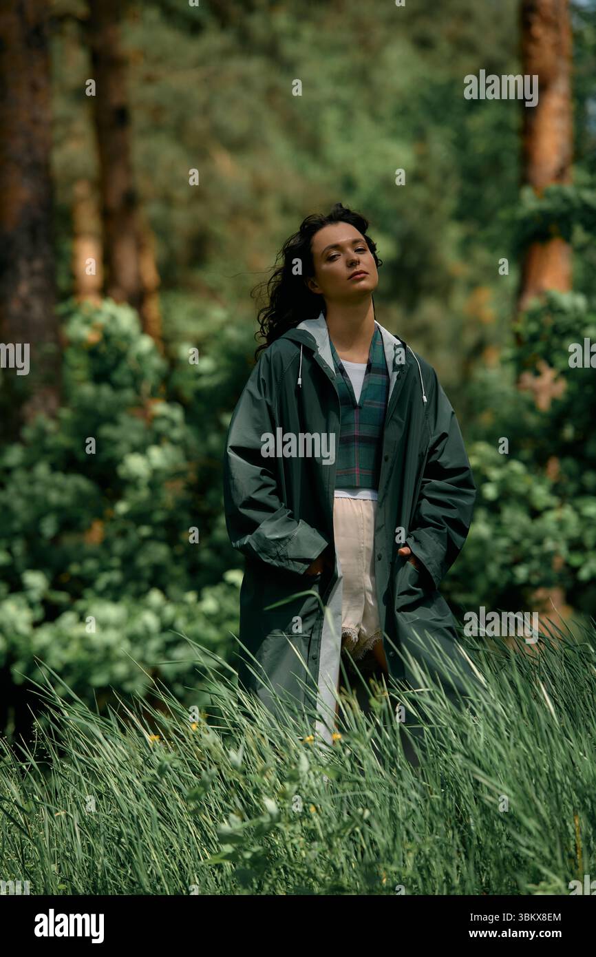 Eine junge Frau mit lockigen Haaren steht selbstbewusst in einem stilvollen Regenmantel inmitten von üppigem Grün. Stockfoto