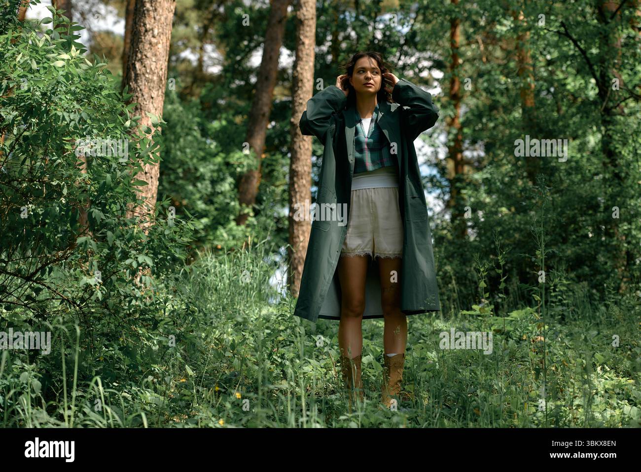 Eine Frau mit lockigen Haaren zeigt ihren modischen Sinn in einem stylischen Regenmantel, während sie draußen im Wald ist. Stockfoto