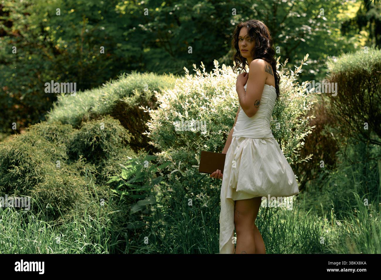 Eine junge Frau mit lockigen Haaren posiert anmutig draußen in einem wunderschönen weißen Kleid. Stockfoto