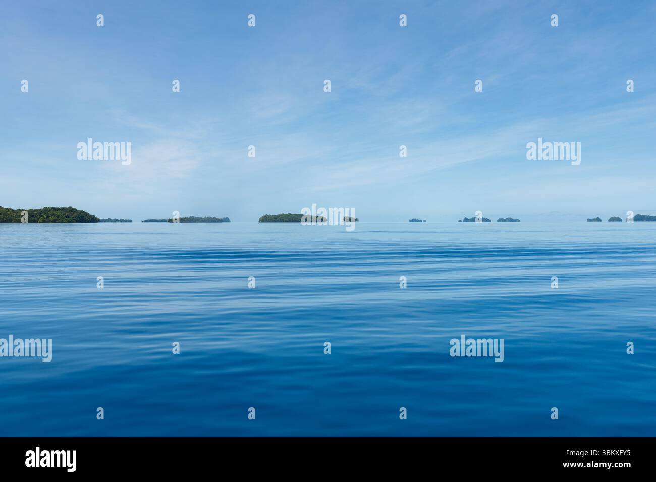 Eine ruhige Meereslandschaft mit ruhigem, blauem Wasser unter einem blassblauen Himmel mit verstreuten Wolken, gesäumt von kleinen, grünen Inseln in der Ferne, die eine ruhige und malerische Szene schaffen. Stockfoto