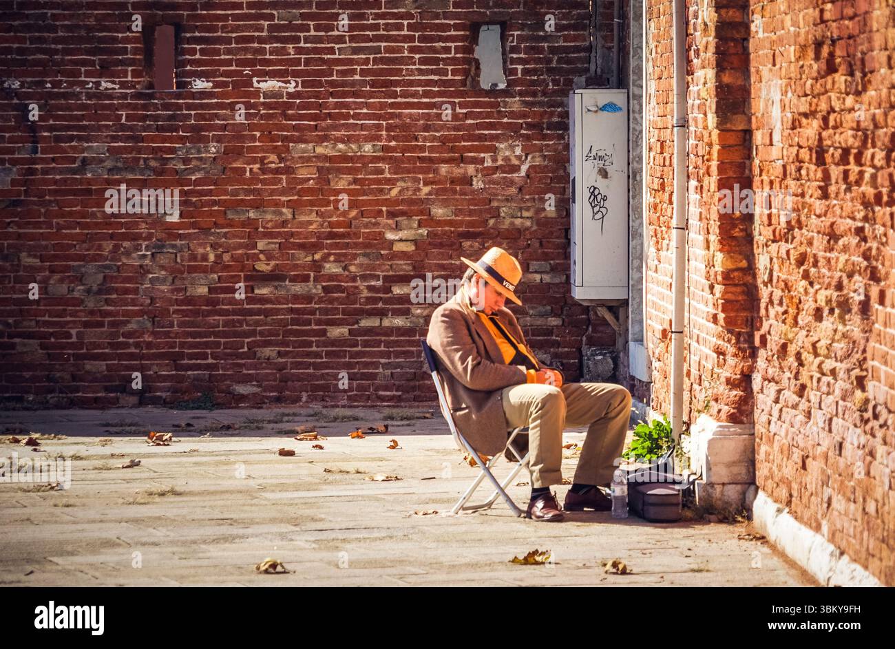 Ein Straßenmusiker rastet in Venedig, Italien, an der verwitterten Backsteinmauer aus - ein authentischer Moment des einheimischen Straßenmusikers an sonnigen Tagen Stockfoto