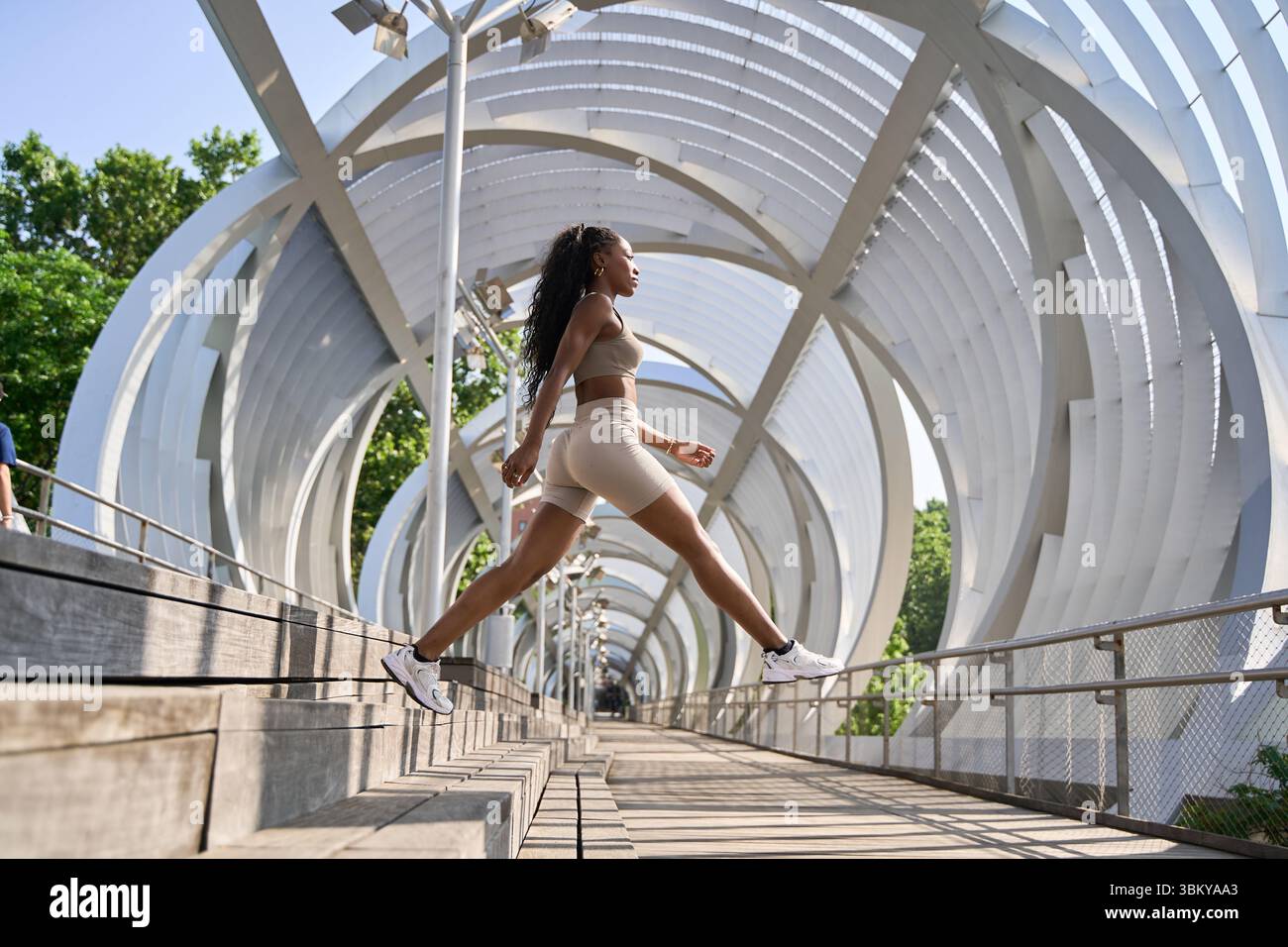 Eine fitte schwarze Frau, die an einem sonnigen Tag Dehnungsübungen macht, springt auf die Treppe einer modernen weißen Metallbrücke in der Stadt Stockfoto