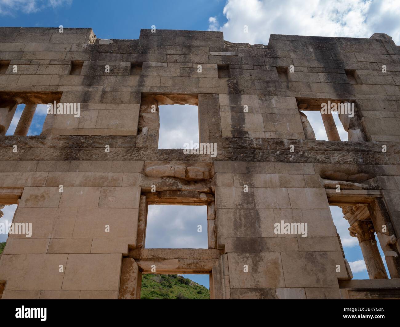Eine Niedrigwinkelaufnahme der Celsus-Bibliothek in Ephesus, Türkei, mit ihrer verwitterten Steinfassade mit rechteckigen Öffnungen, die einst Fenster oder architektonische Details enthielten, vor dem Hintergrund eines teilweise bewölkten Himmels und fernen Grüns, die die historische Bedeutung und das architektonische Design der antiken Struktur hervorheben. Stockfoto