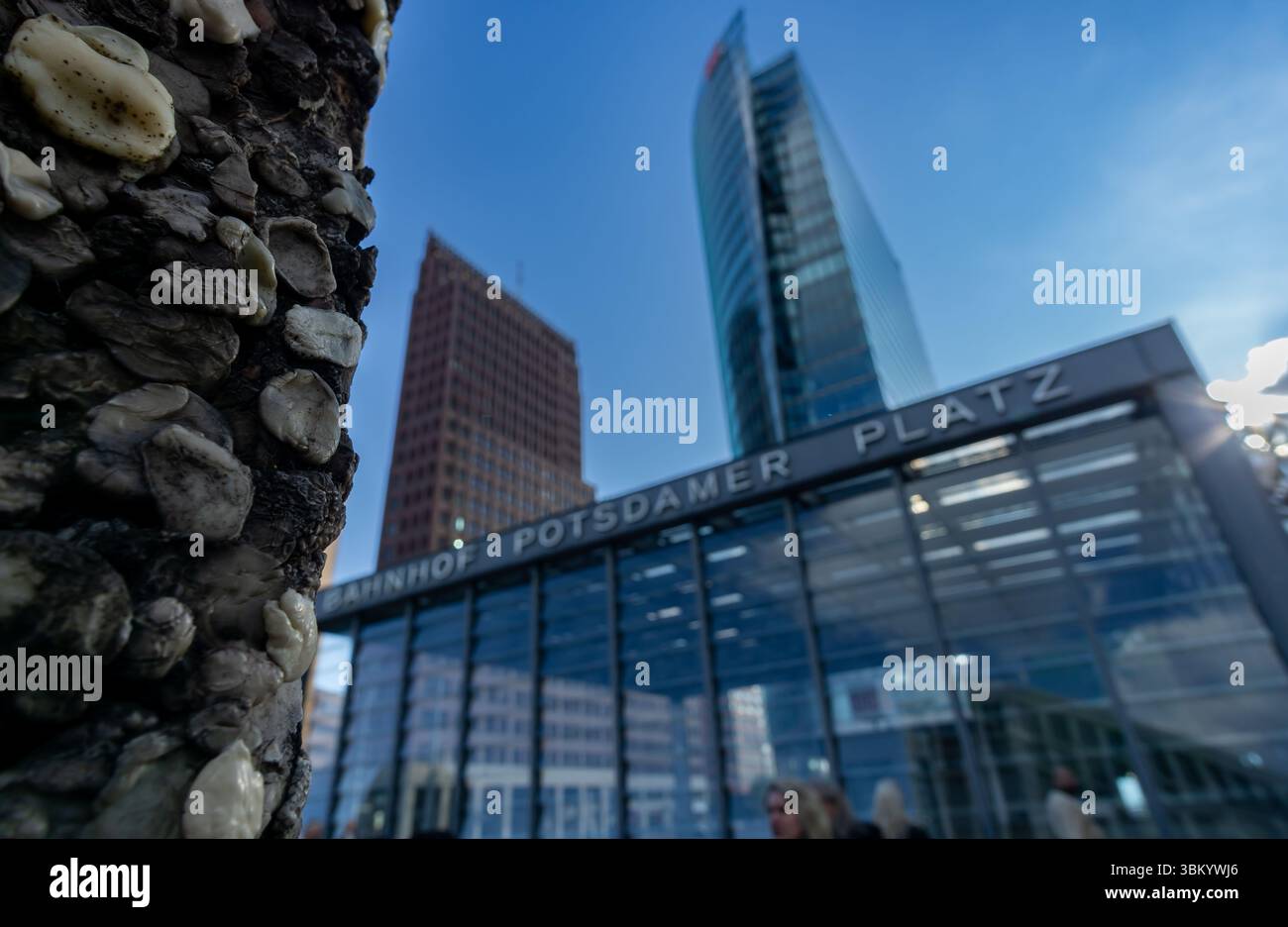 Scharfer Fokus auf ein Berliner Mauer-Segment mit verschwommenem Potsdamer Platz und urbaner Menschenmenge im Hintergrund. Stockfoto