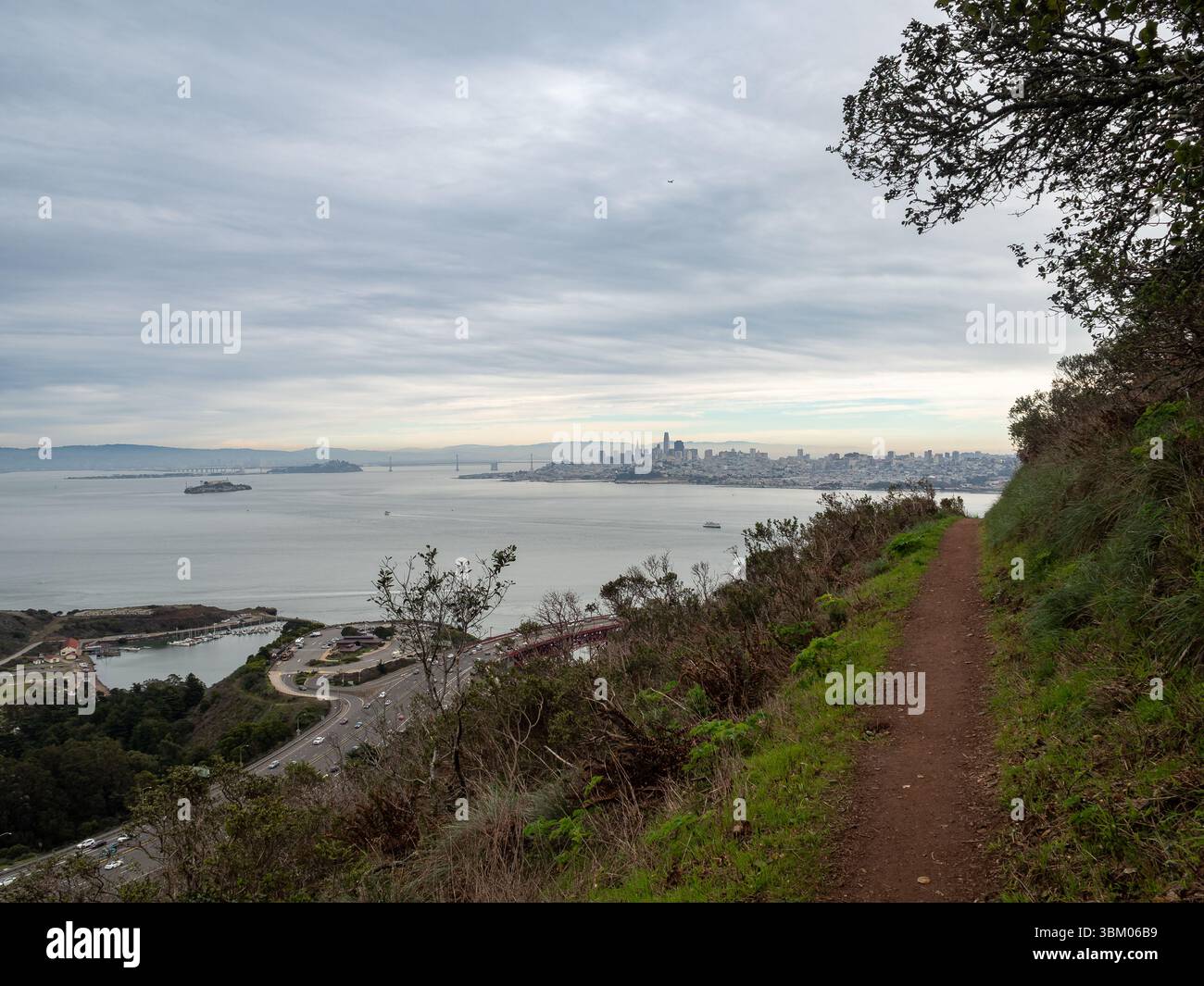 Ein malerischer Blick von einem Wanderweg mit Blick auf die San Francisco Bay, Alcatraz Island, die Golden Gate Bridge und die Skyline von San Francisco unter einem bewölkten Himmel, die die natürliche Schönheit und urbane Landschaft der Bay Area zeigt. Stockfoto